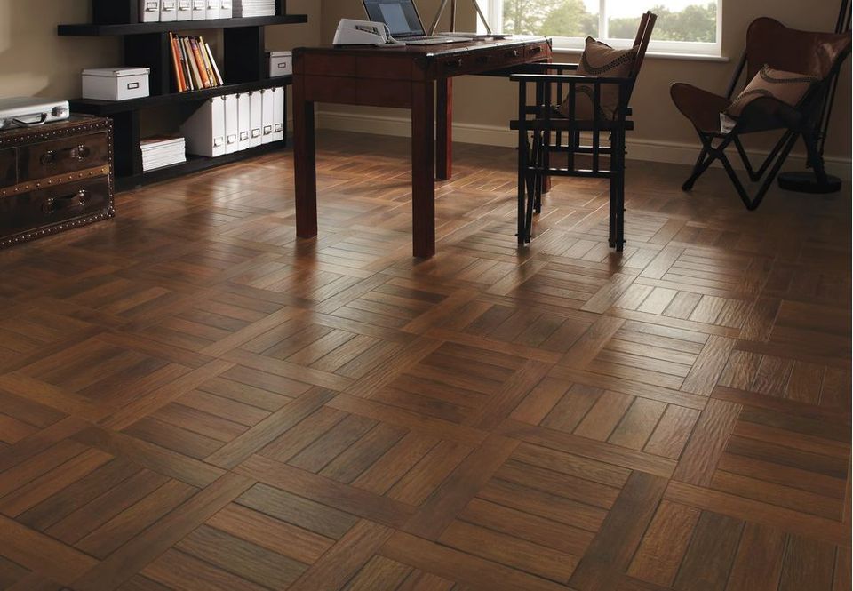 The image shows a spacious room with wooden flooring. The flooring has a natural wood color and texture, with visible knots and grain patterns that give it a rustic appearance. The planks of flooring are laid out in a traditional pattern, with a subtle shine that reflects the natural light coming in from the large windows in the room. The wooden flooring complements the overall aesthetic of the room, which appears to be a mix of modern and traditional elements.